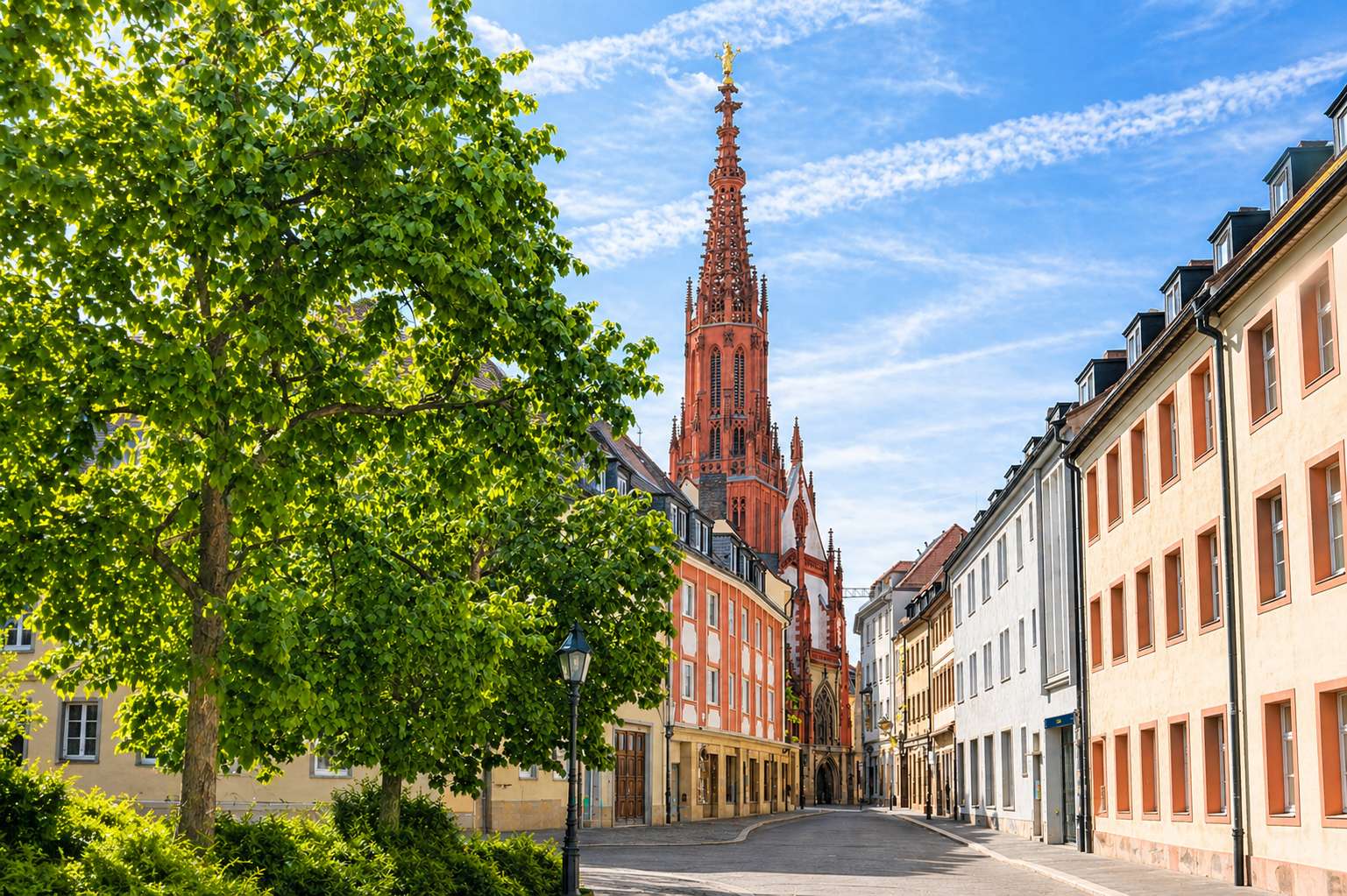 Ruhige Altstadtstraße in Würzburg mit hellen Häuserfassaden und der hohen roten gotischen Kirchturmspitze der Marienkapelle unter blauem Himmel, eingerahmt von grünen Bäumen.