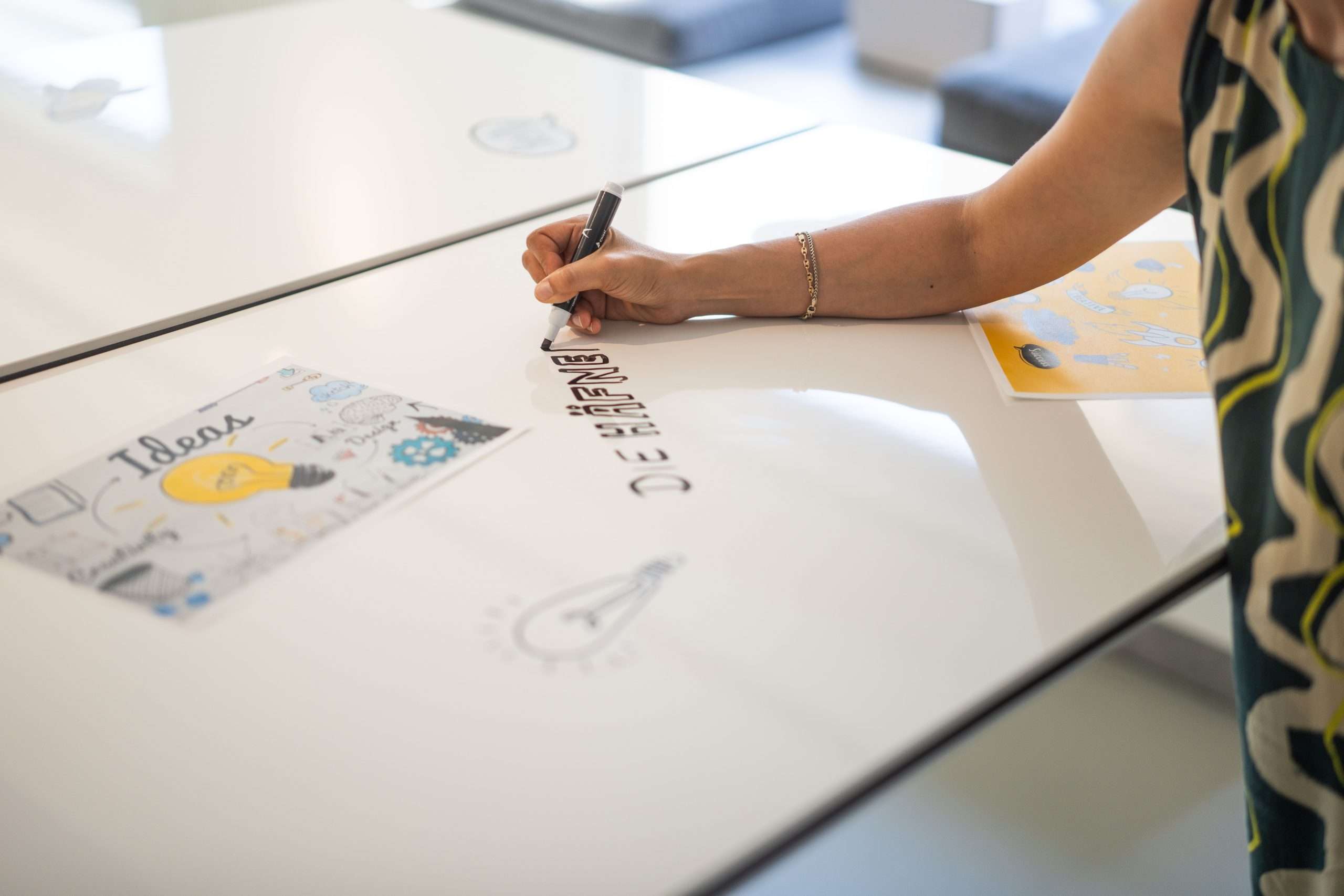 Hand einer Workshop-Teilnehmerin, die bei Die Häfnerei in Würzburg Ideen auf einen beschreibbaren Tisch schreibt, umgeben von Skizzen und Visualisierungen.